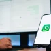 Celular exibindo o logo do WhatsApp em frente a dois monitores de computador. A tela de fundo exibe uma interface de chat do WhatsApp.