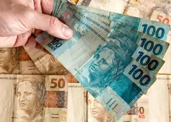 Notas de cem e cinquenta reais sendo seguradas por uma mão, representando calendário de pagamentos do INSS.