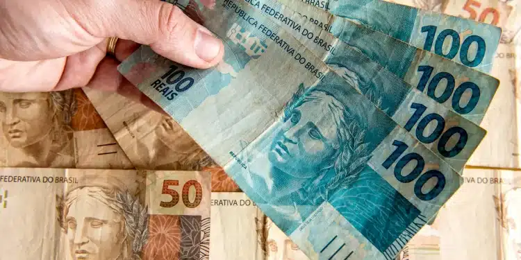 Notas de cem e cinquenta reais sendo seguradas por uma mão, representando calendário de pagamentos do INSS.