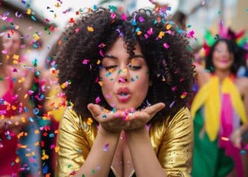 Mulher jovem de cabelos cacheados e jaqueta dourada soprando confetes coloridos em uma festa de rua animada durante o Carnaval.