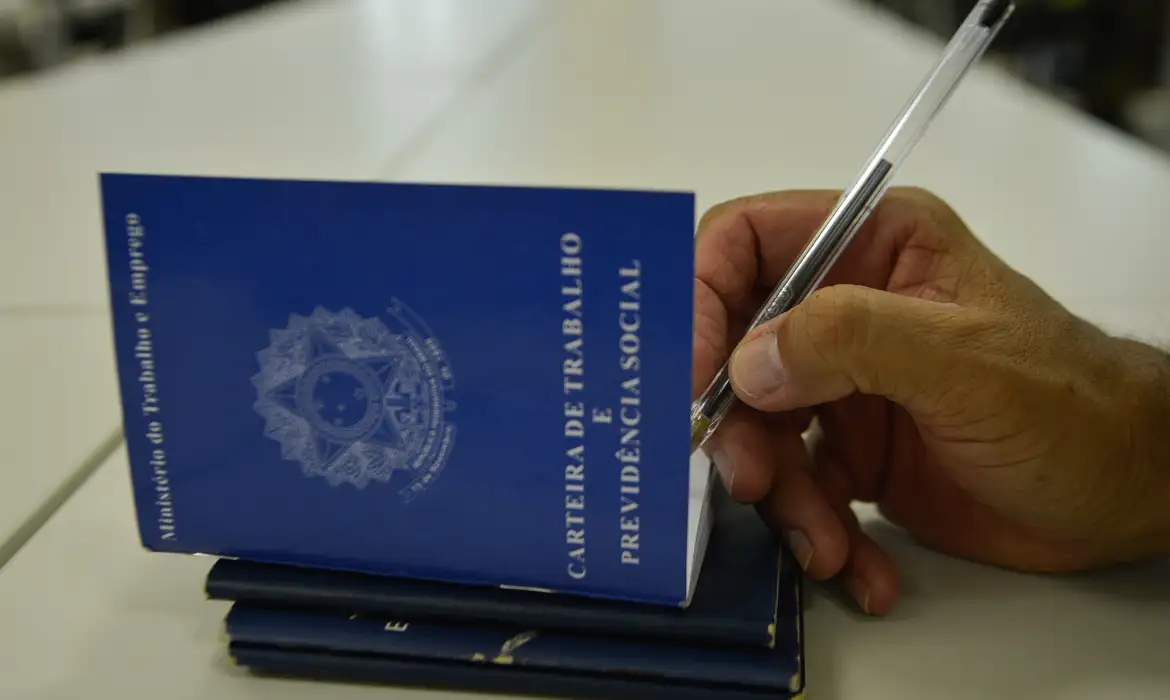 Mão de pessoa escreve com caneta em Carteira de Trabalho azul sobre mesa