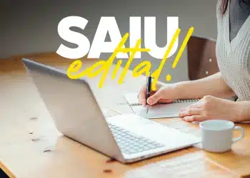 Pessoa escrevendo em caderno ao lado de notebook com destaque para anúncio de edital