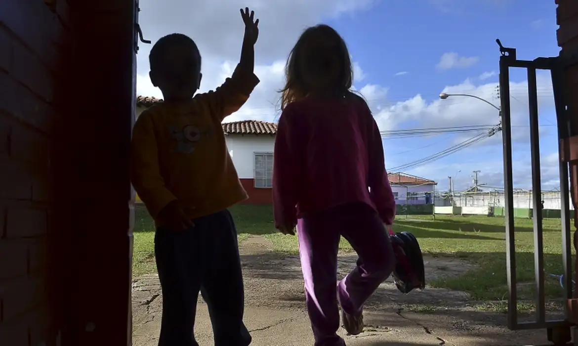 Duas crianças de costas brincam próximas a portão aberto com céu azul ao fundo