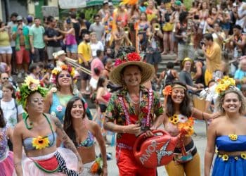 Close-up de um calendário de mesa aberto no mês de fevereiro de 2027. Uma mão segura uma caneta vermelha e circula os dias 6, 7, 8 e 9 (Sábado a Terça de Carnaval), com confetes coloridos e serpentinas espalhados sobre a mesa ao redor do calendário.