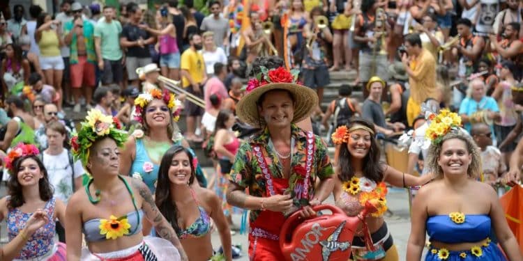 Close-up de um calendário de mesa aberto no mês de fevereiro de 2027. Uma mão segura uma caneta vermelha e circula os dias 6, 7, 8 e 9 (Sábado a Terça de Carnaval), com confetes coloridos e serpentinas espalhados sobre a mesa ao redor do calendário.