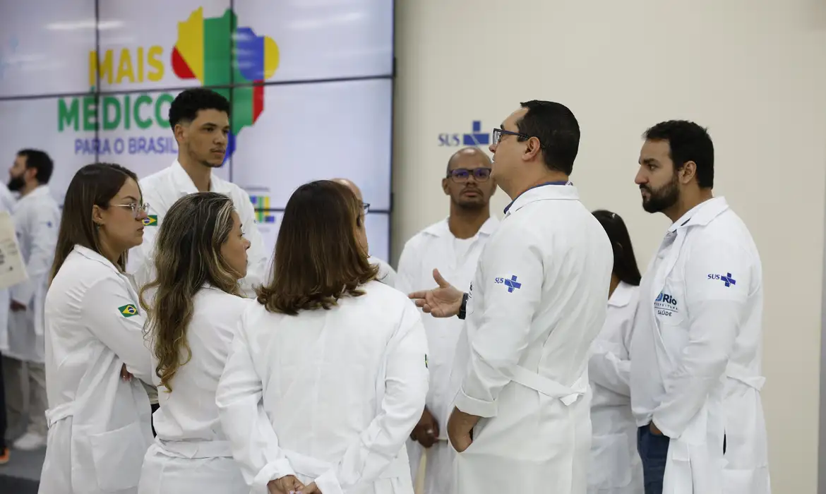 Grupo de médicos em jaleco branco conversa em frente a painel do programa Mais Médicos
