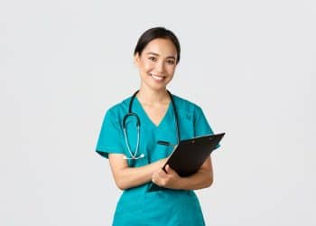 Uma enfermeira sorridente de ascendência asiática veste um uniforme cirúrgico (scrub) na cor azul-petróleo e um estetoscópio preto em volta do pescoço. Ela segura uma prancheta preta com a mão esquerda e uma caneta com a direita, posicionada como se estivesse fazendo anotações, contra um fundo branco e limpo.