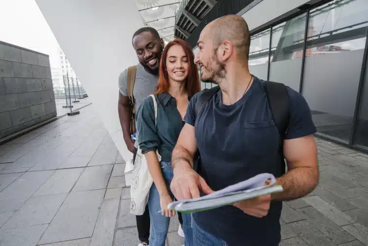 Três estudantes conversam sorrindo enquanto revisam anotações ao ar livre em frente a prédio universitário