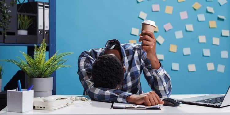 Um trabalhador de pele escura com cabelo crespo, vestindo camisa xadrez azul e branca, está com a cabeça abaixada sobre a mesa em um escritório moderno de fundo azul com post-its. Ele segura um copo de café descartável, visivelmente exausto ao lado de um notebook e um telefone fixo.