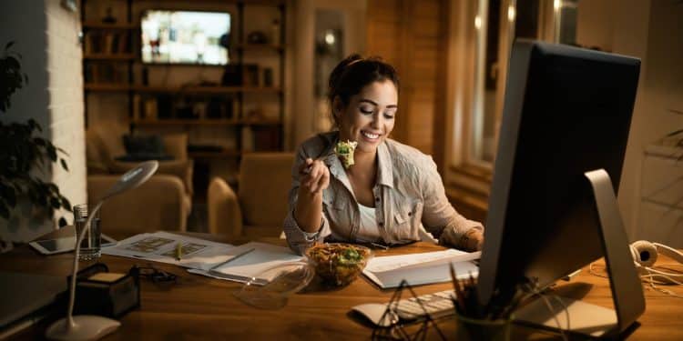 Mulher sorrindo enquanto come salada e trabalha em computador de mesa durante expediente noturno em home office