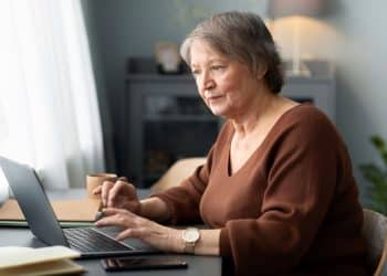 Mulher sênior de cabelos curtos grisalhos, vestindo uma blusa marrom, sentada à mesa e utilizando um notebook com foco e atenção em um ambiente doméstico iluminado.