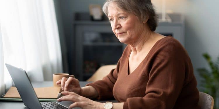 Mulher sênior de cabelos curtos grisalhos, vestindo uma blusa marrom, sentada à mesa e utilizando um notebook com foco e atenção em um ambiente doméstico iluminado.