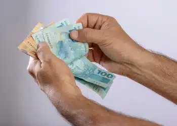 Pessoa segurando cédulas de dinheiro brasileiras, destacando o valor de R$ 100 e R$ 50, representando a movimentação do Pé-de-Meia.