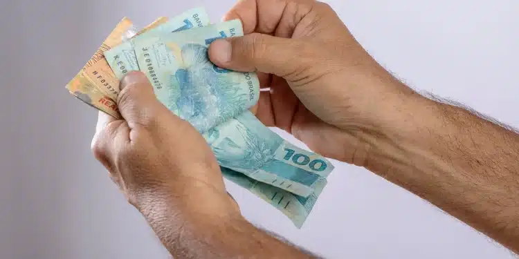 Pessoa segurando cédulas de dinheiro brasileiras, destacando o valor de R$ 100 e R$ 50, representando a movimentação do Pé-de-Meia.