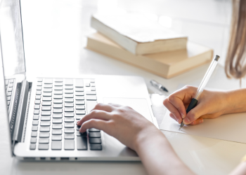 Pessoa escrevendo em uma folha enquanto usa um laptop para inscrição em concurso.