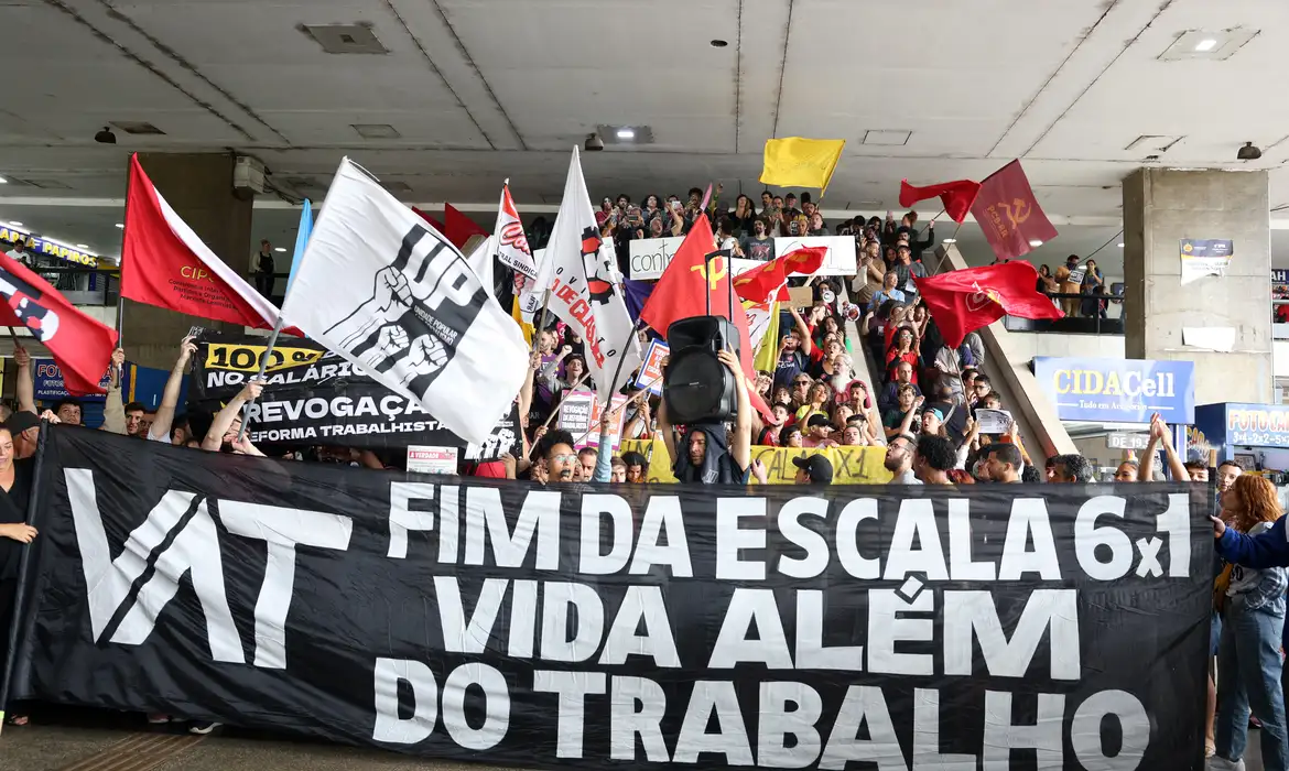 Manifestantes em protesto contra a jornada de trabalho 6x1, com bandeiras e um grande banner exigindo uma mudança nas condições de trabalho no Brasil.