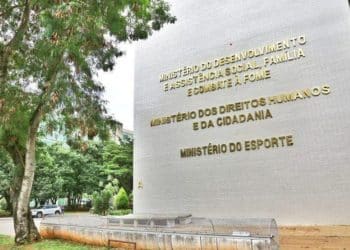 Vista lateral da fachada de um edifício governamental em Brasília, com paredes brancas e letras douradas que identificam o Ministério do Desenvolvimento Social, Família e Combate à Fome, o Ministério dos Direitos Humanos e o Ministério do Esporte. À esquerda, árvores verdes e um carro estacionado compõem o cenário sob luz natural.