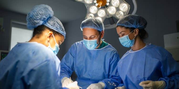 Três cirurgiões, dois homens e uma mulher, realizam um procedimento médico em um centro cirúrgico iluminado. Eles vestem uniformes azuis, máscaras, toucas e luvas descartáveis, focados no trabalho sob a luz de um foco cirúrgico de LED no teto.