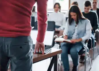 Professora em sala dirige-se a alunos atentos enquanto uma aluna anota conteúdos em caderno
