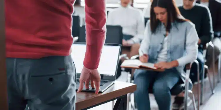 Professora em sala dirige-se a alunos atentos enquanto uma aluna anota conteúdos em caderno