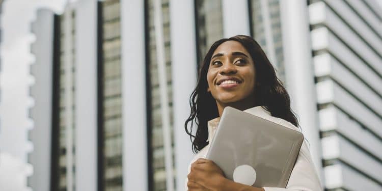 Mulher negra jovem e sorridente, vestindo camisa branca, segurando um notebook fechado contra o peito e olhando para o horizonte. Ao fundo, prédios comerciais modernos em tons de cinza e branco sob um céu claro.