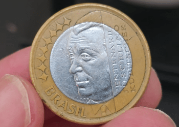 Close detalhado da moeda de 1 real JK sendo segurada por dedos. A face prateada exibe o perfil de Juscelino Kubitschek com a inscrição "CENTENÁRIO JUSCELINO KUBITSCHEK", enquanto o anel dourado externo mostra a palavra "BRASIL" e estrelas decorativas.