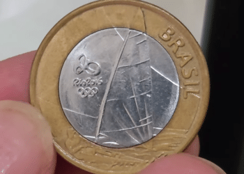 Close-up de uma moeda de 1 real das Olimpíadas Rio 2016 (tema vela), sendo segurada por dedos, mostrando a borda dourada e o centro prateado com o desenho do barco.