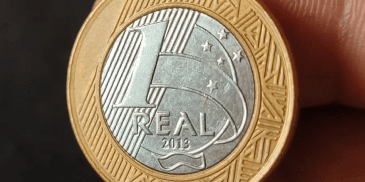 Close de uma moeda de 1 real do ano de 2013 sendo segurada pelos dedos, mostrando detalhadamente o valor de face e os elementos de segurança no reverso prateado.