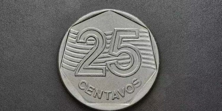Vista em close-up da face anversa de uma moeda de 25 centavos de real, mostrando o valor numérico "25" e a palavra "CENTAVOS" em relevo, em fundo escuro e liso.