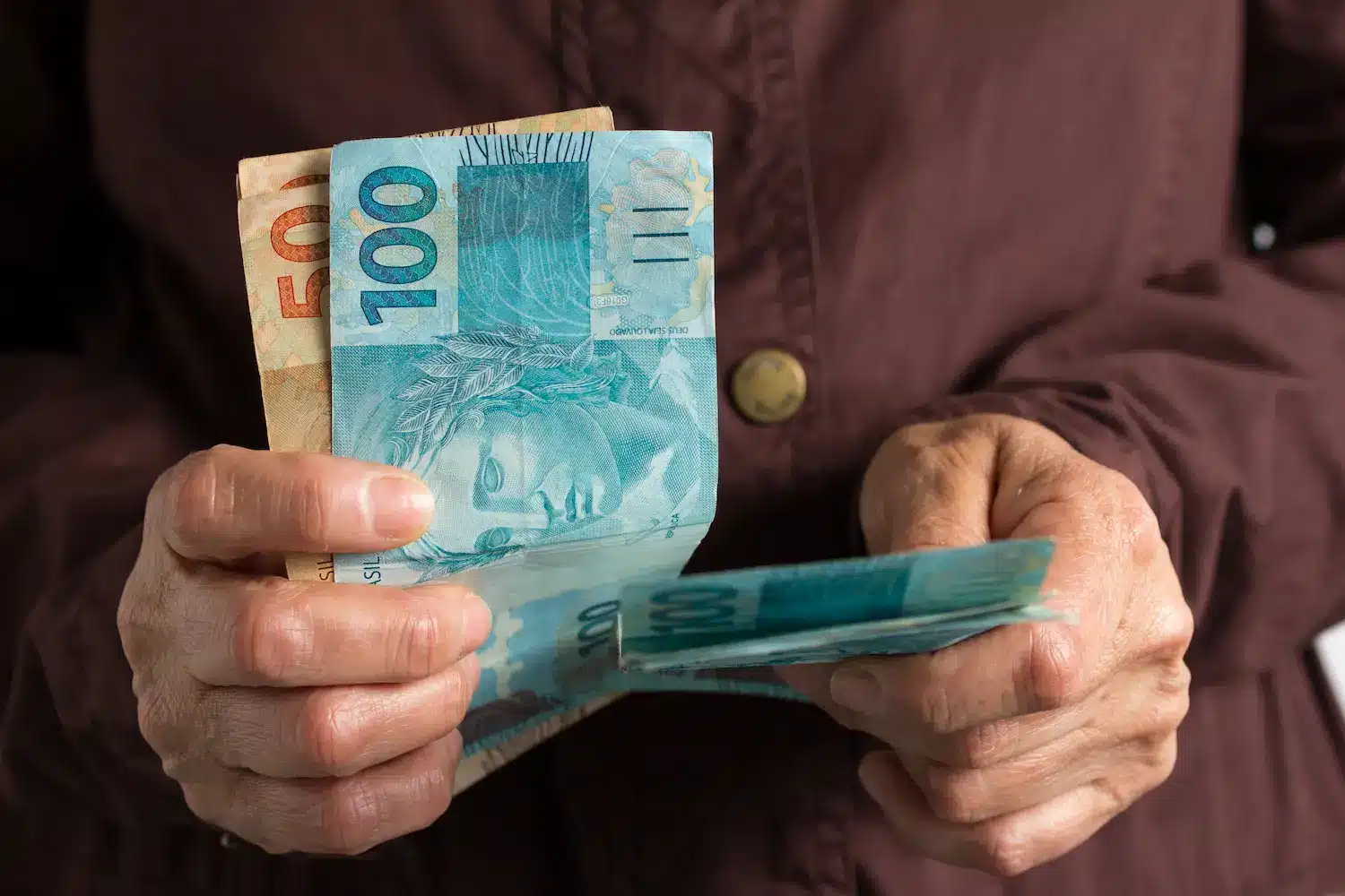 Mãos segurando notas de 100 reais ilustram as regras para liberação imediata de R$ 1.000 do programa Pé-de-Meia.