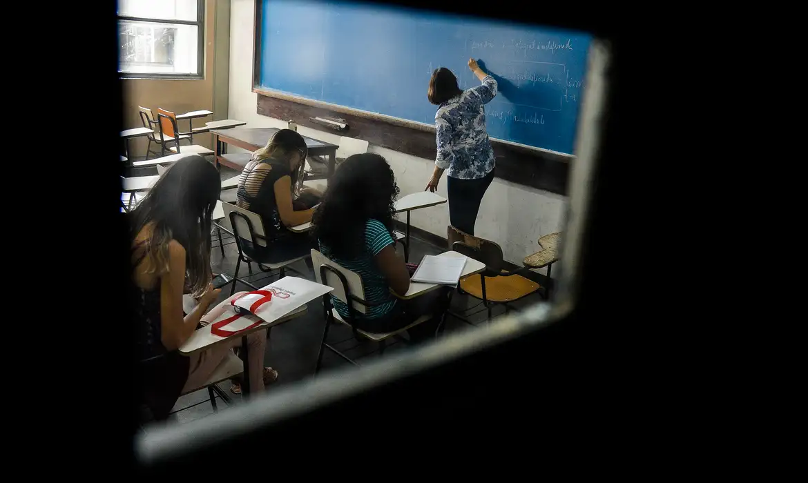 Estudantes sentados escrevendo enquanto professora bolsista do Pé-de-Meia Licenciaturas escreve em quadro azul de sala iluminada