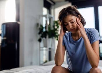 Mulher jovem de camiseta azul sentada na beira da cama, com os olhos fechados e as mãos pressionando as têmporas em sinal de cansaço ou dor de cabeça.