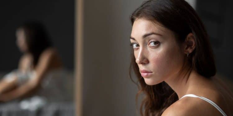 Retrato em close de uma mulher jovem de cabelos castanhos ondulados e olhar firme e direto, transmitindo compostura e serenidade. Ao fundo, seu reflexo em um espelho aparece levemente desfocado, simbolizando a introspecção e o domínio próprio. Ela veste uma regata branca simples em um ambiente de tons neutros.