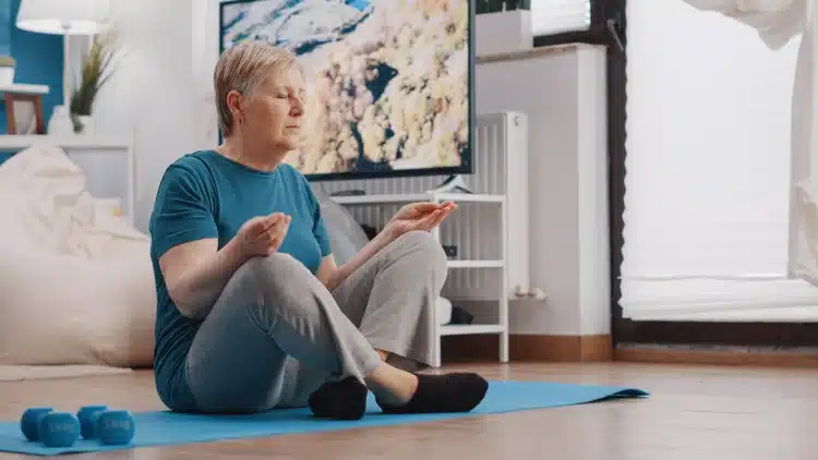 Idosa sentada no tapete azul meditando com olhos fechados e expressão serena