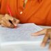 Pessoa escrevendo com caneta vermelha em caderno quadriculado durante prova ou estudo