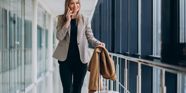 Profissional elegante falando ao celular em corredor de aeroporto com mala de viagem e casaco no braço representando carreiras que permitem viajar