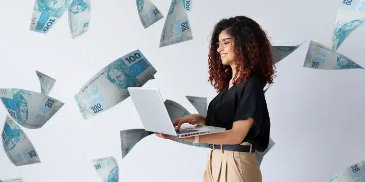 Mulher sorrindo enquanto usa laptop, com notas de 100 reais flutuando ao seu redor, representando o sucesso financeiro e o trabalho remoto.