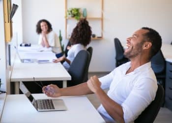 Homem sorrindo e segurando óculos, trabalhando em um escritório moderno com colegas de trabalho ao fundo.