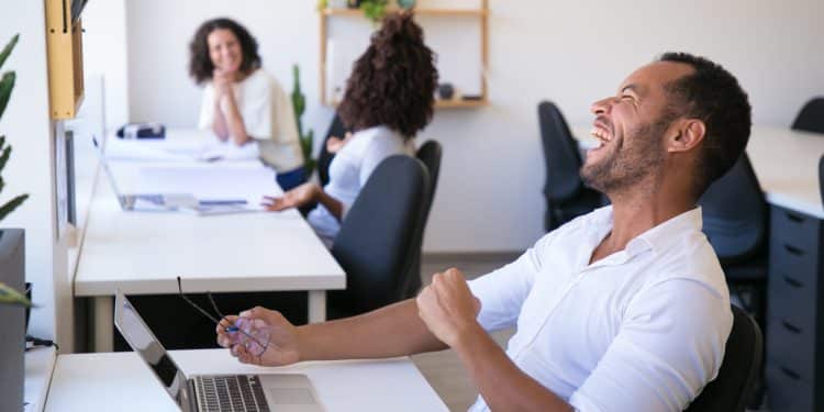Homem sorrindo e segurando óculos, trabalhando em um escritório moderno com colegas de trabalho ao fundo.
