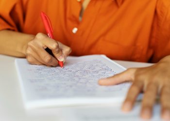 Pessoa escrevendo com caneta vermelha em caderno quadriculado representando candidato estudando os salários e benefícios do concurso SAMAE Campos Novos SC