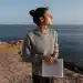 Mulher em momento de introspecção à beira-mar segurando um livro aberto durante o pôr do sol.