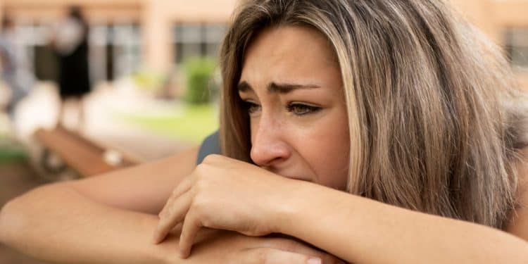 Mulher jovem com expressão triste e pensativa, apoiando o rosto sobre as mãos em um banco de madeira ao ar livre, representando o peso emocional da culpa.