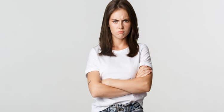 Uma mulher jovem de camiseta branca e calça jeans, com os braços cruzados e uma expressão facial de descontentamento e irritação contra um fundo branco.