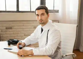 Homem médico em consultório, com estetoscópio no pescoço, segurando um dispositivo e olhando diretamente para a câmera.
