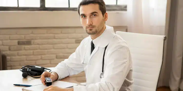 Homem médico em consultório, com estetoscópio no pescoço, segurando um dispositivo e olhando diretamente para a câmera.