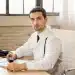 Homem médico em consultório, com estetoscópio no pescoço, segurando um dispositivo e olhando diretamente para a câmera.