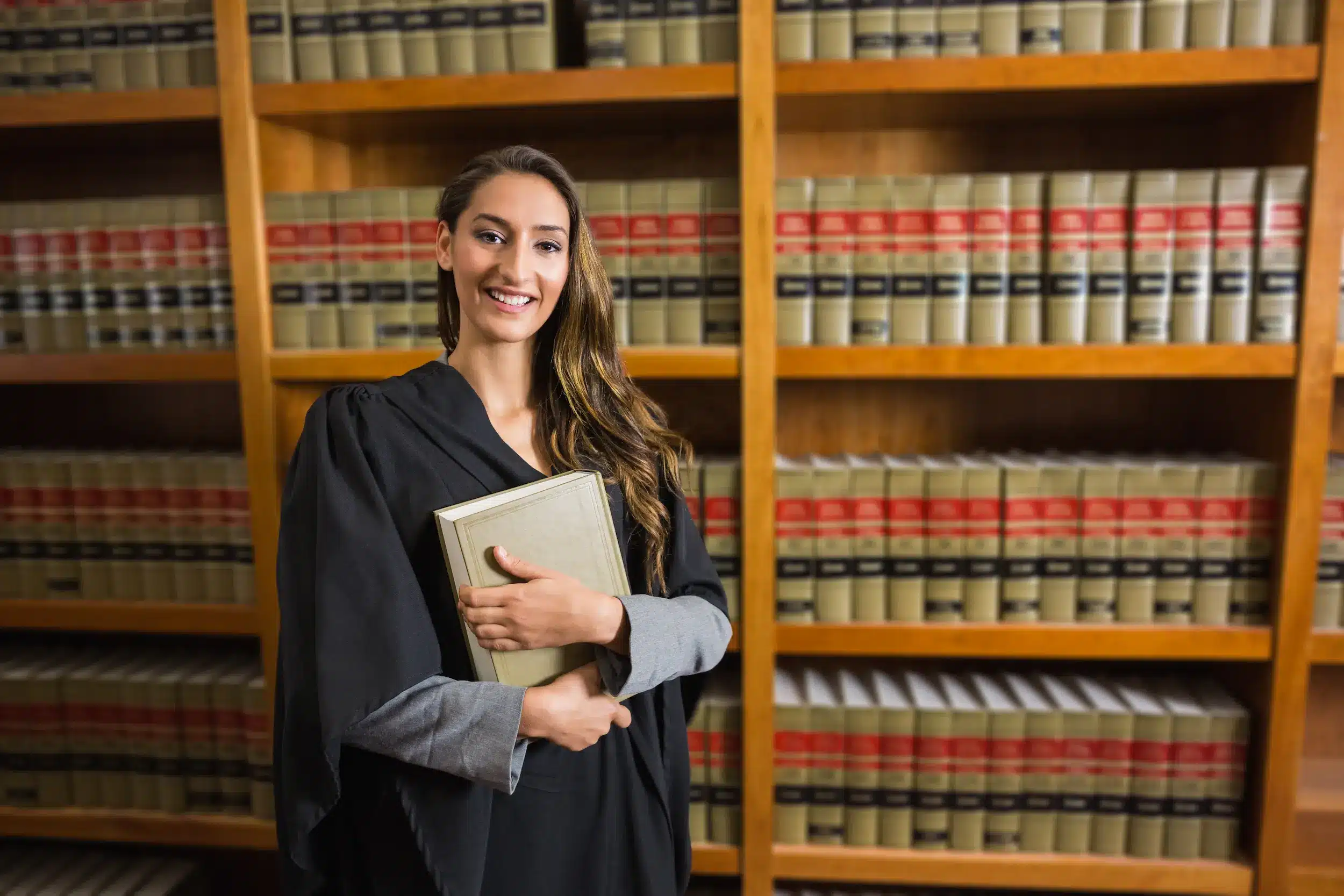 Mulher advogada sorrindo em frente a uma estante de livros jurídicos, segurando um livro com as duas mãos.