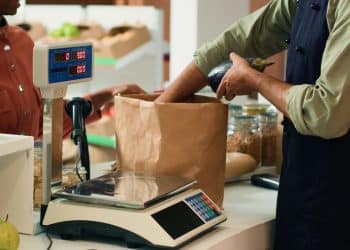 Funcionário pesando produto em balança digital no comércio, enquanto outro funcionário prepara sacola de papel, em contexto de atendimento ao cliente.