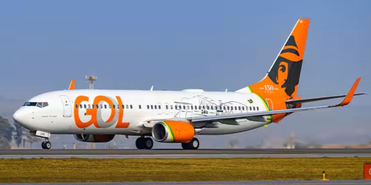 Aeronave Boeing 737 da GOL Linhas Aéreas taxiando em uma pista de aeroporto sob o sol. O avião possui a fuselagem branca com o logotipo laranja e uma pintura especial na cauda celebrando 150 anos com a imagem de Santos Dumont.