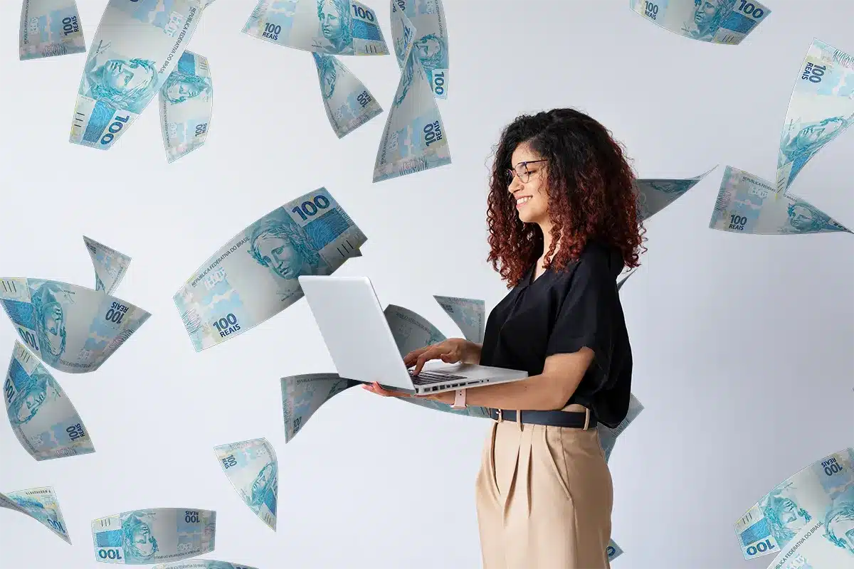 Mulher sorridente usando um notebook enquanto notas de 100 reais flutuam ao redor, simbolizando o aumento salarial feminino em algumas profissões.